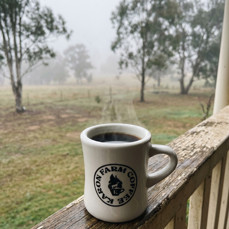 Mug of coffee with Karon Farm Coffee logo on a wooden railing overlooking a misty field.